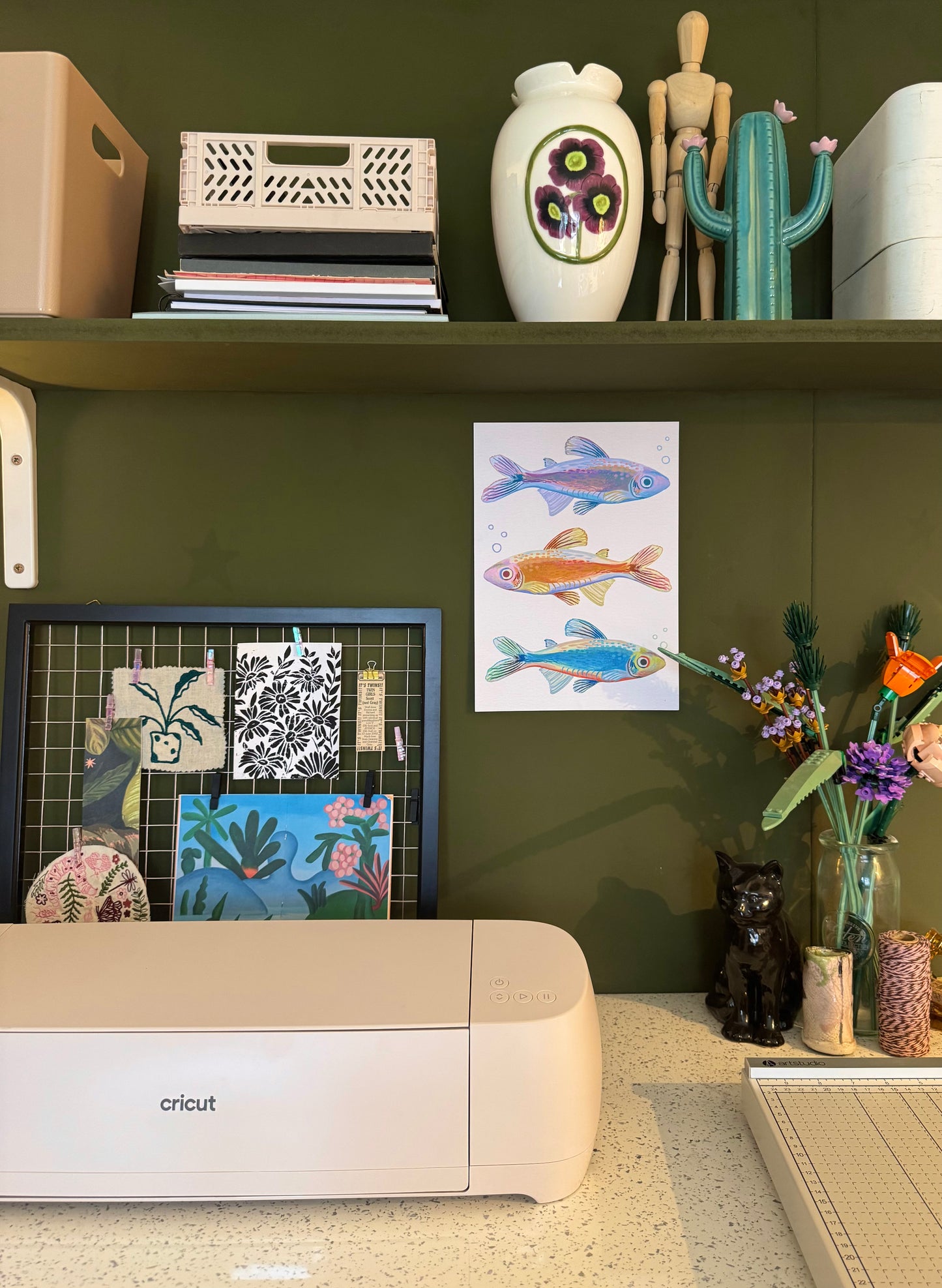 Image of studio focusing on green wall, shelf with decorative items, and the framed artwork up on the wall.