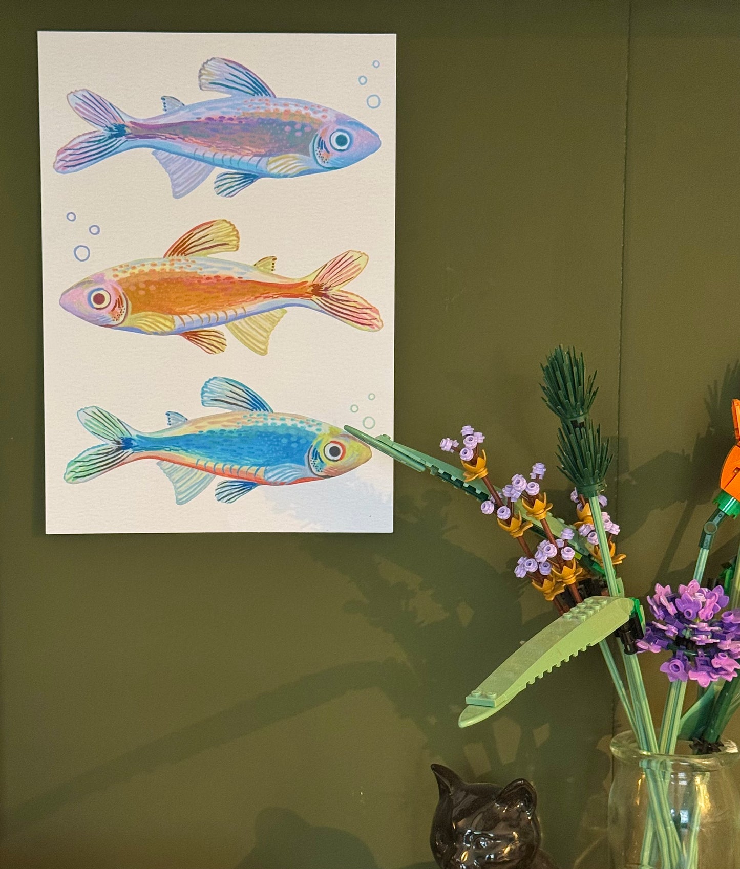 Colorful fish illustration on a white sheet next to a vase with flowers on a green wall.